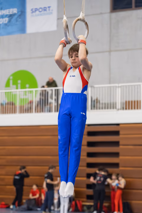 Young gymnast in red, white and blue uniform performs still rings routine with focused determination in indoor gymnasium