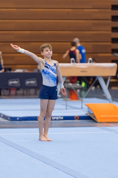 Young gymnast performing floor routine with one arm extended, focused expression, blue leotard, gymnasium setting