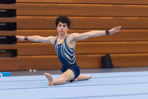 Male gymnast performs floor routine in split position with arms extended, wearing teal leotard in indoor training facility