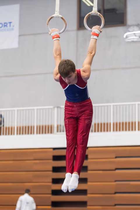 Young gymnast performs a still rings hold, arms extended overhead, wearing maroon and blue uniform in indoor gymnasium