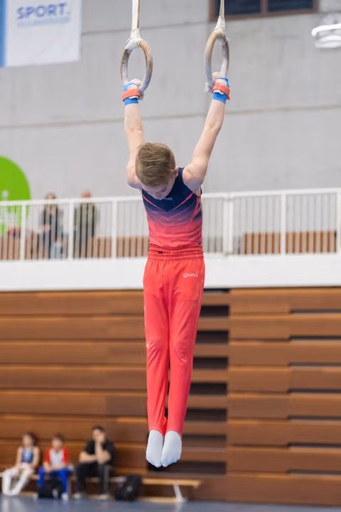 Young gymnast in gradient pink outfit hangs from rings with extended arms during routine in indoor gymnasium