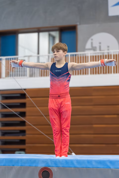 Young gymnast stands on balance beam with arms extended horizontally, wearing pink gradient leotard in training facility
