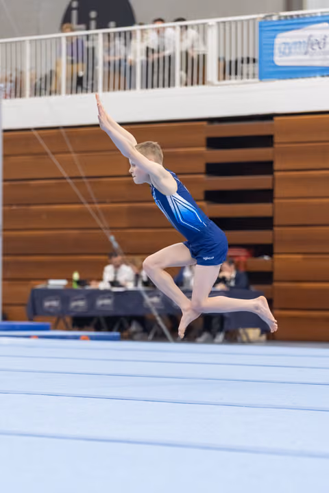 Gymnast in blue leotard performs a dynamic leap with extended arms during floor exercise routine in indoor gymnasium