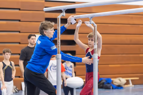 Young gymnast grips parallel bars while coach in blue adjusts equipment, wooden bleachers in background
