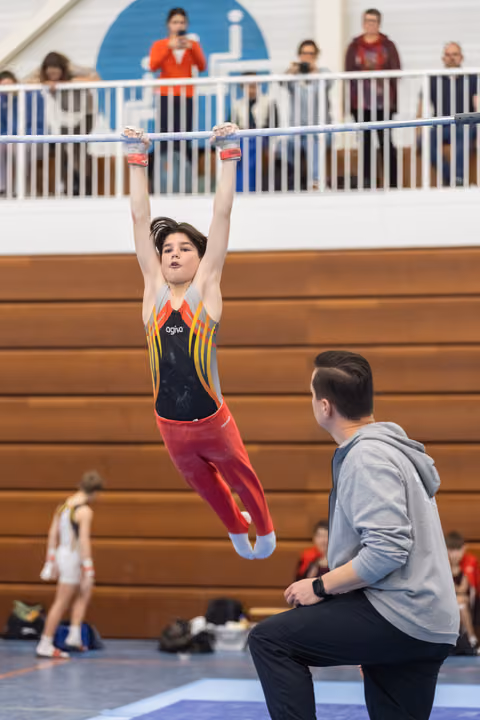 Young gymnast in colorful leotard performs a hanging routine on the bar while coach observes closely below