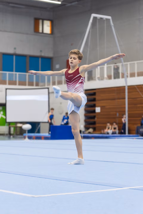 Young gymnast performing a split leap during floor routine at indoor gym, arms extended with focused expression