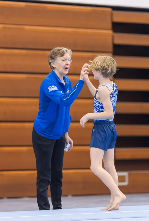 Coach in blue jacket offers guidance and encouragement to young gymnast in blue leotard during floor exercise training