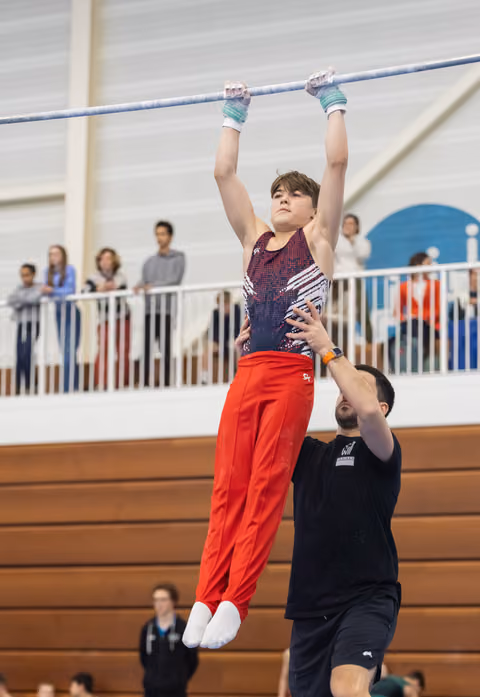 Young gymnast in red pants and sparkly leotard hangs from high bar while coach spots from below in indoor gymnasium