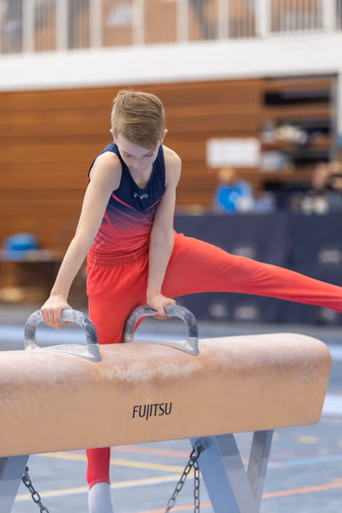 Young gymnast in red outfit performs on pommel horse with focused concentration during training session