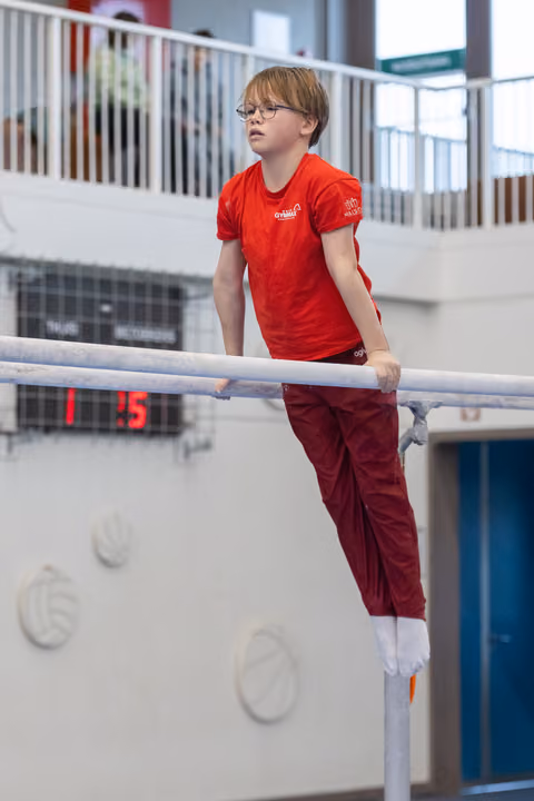 Young gymnast in red shirt and glasses performs on parallel bars, displaying focus and determination in training facility