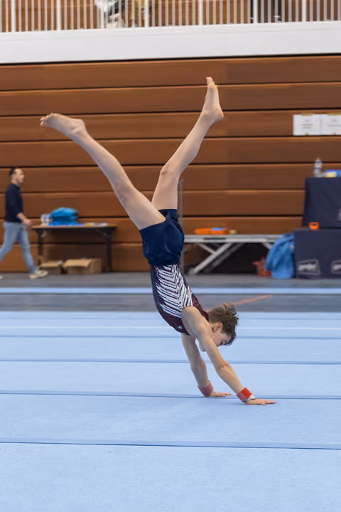 Young gymnast performing a handstand on blue floor mat, legs extended upward, in training facility