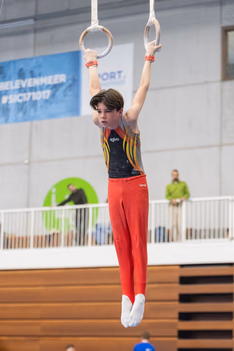 Young gymnast in red pants and striped leotard hangs from rings with arms extended, showing focused concentration