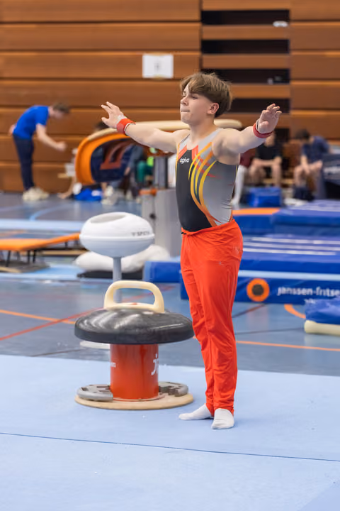 Young gymnast in red pants and gray leotard strikes a pose with arms extended beside a mushroom circle apparatus