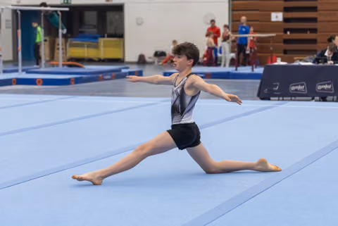 Young gymnast executes a graceful split leap during floor routine, arms extended, in front of spectators and judges
