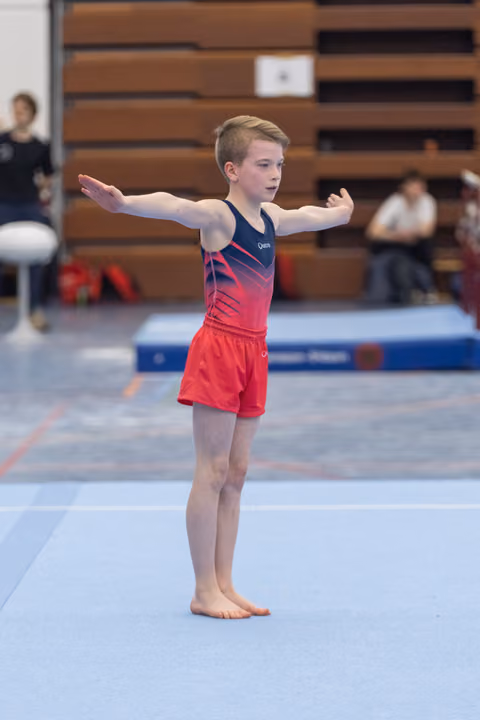Young male gymnast performs floor routine with arms extended, wearing red gradient leotard in gymnasium