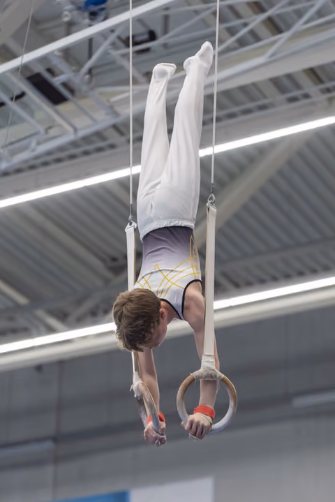 Young male gymnast performing an inverted hold on still rings in an indoor training facility, demonstrating strength and control