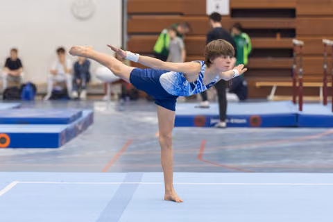 Young gymnast performs a T-pose balance on floor, arms extended horizontally, demonstrating flexibility and control