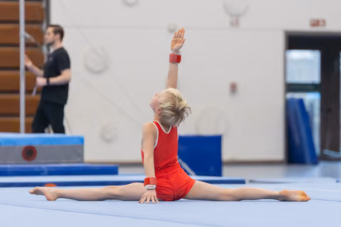Young gymnast in red leotard performs a full split on floor mat with one arm raised, demonstrating flexibility