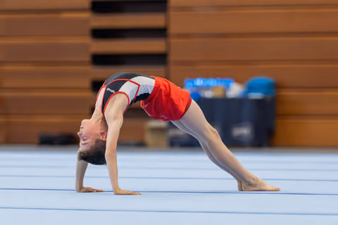 Young gymnast performs a back bridge on the floor mat, smiling upward with visible flexibility and confidence.