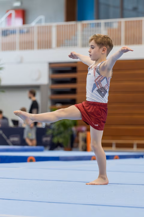 Young male gymnast performs an arabesque on the floor exercise mat, arms extended with focused expression.