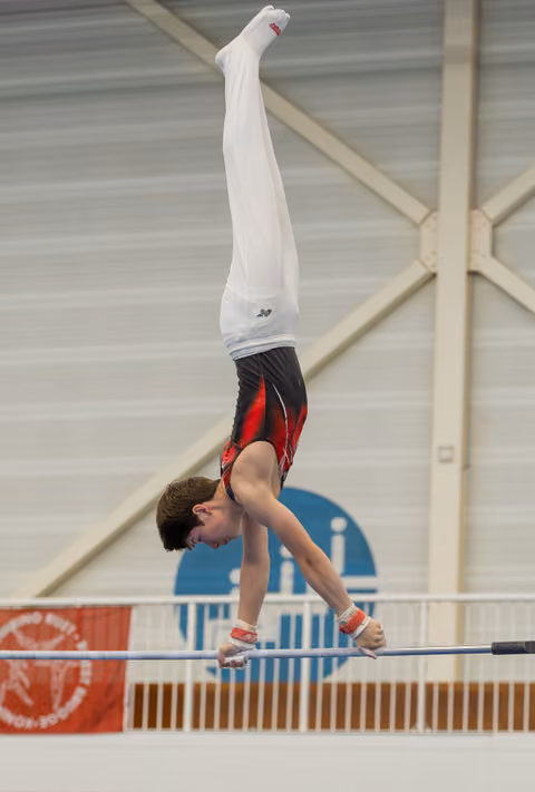 Young male gymnast performs a perfect handstand on the high bar, body fully extended, in an indoor gymnastics hall.