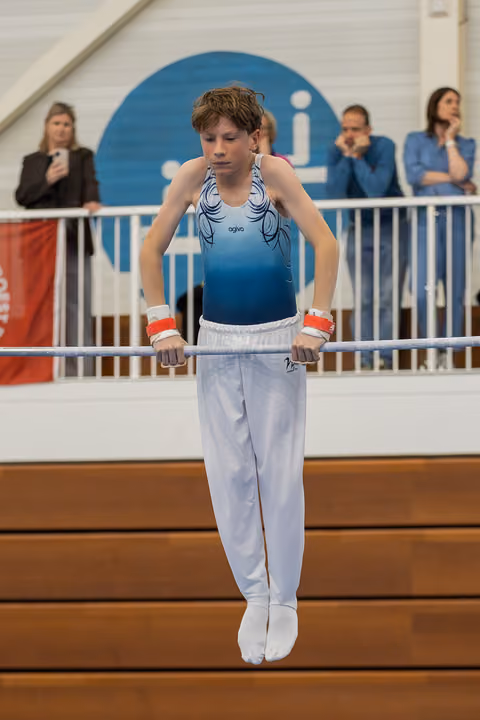 Young male gymnast performing on parallel bars, gripping tightly with focused expression, wearing blue leotard and white trousers.
