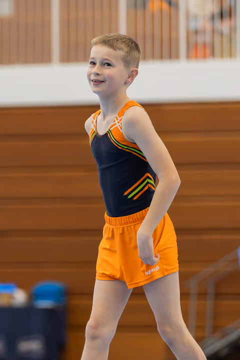 Young male gymnast smiles confidently in orange shorts and navy leotard, standing relaxed on the gymnastics floor.