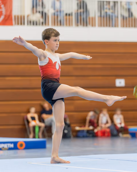Focused young male gymnast holds a one-legged arabesque pose, arms extended, performing on the floor in a sports hall.