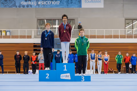 Three young gymnasts stand proudly on a Gymfed podium, gold medalist centered, all wearing medals in a Belgian sports hall.