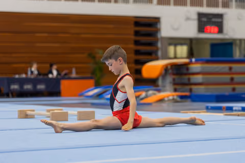 Young boy gymnast performs a perfect split on the blue floor mat, looking down with intense concentration during a meet.