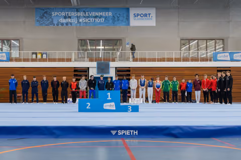 Young gymnasts line up beside a 1-2-3 podium inside a Sport Vlaanderen gymnastics hall, posing formally for awards.