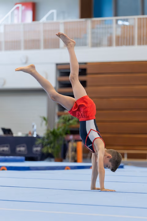 Young male gymnast performs a handstand with legs split apart on the floor exercise mat in an indoor gymnastics hall.