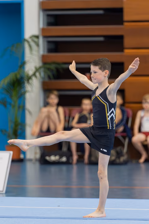 Focused young boy performs a precise arabesque on the gymnastics floor, arms extended, one leg raised high.