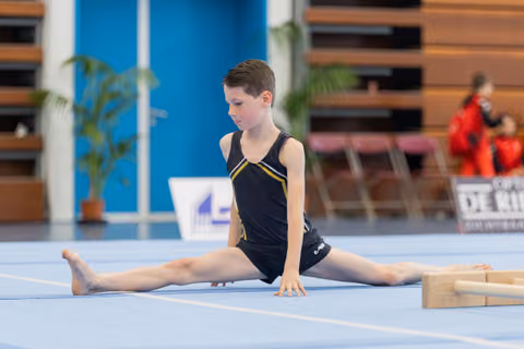 Young boy gymnast performs a perfect split on blue floor mat, focused expression, hands pressed to ground.