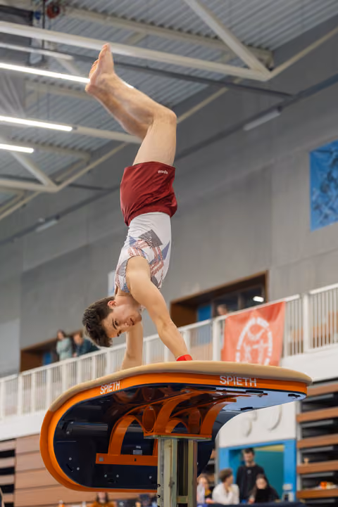 Male gymnast holds a precise handstand on a Spieth vault, legs extended upward, in a packed indoor gymnasium.