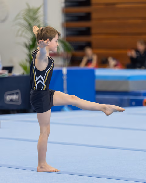 Young male gymnast performs a precise leg raise on the floor exercise mat, arm raised, expression calm and concentrated.