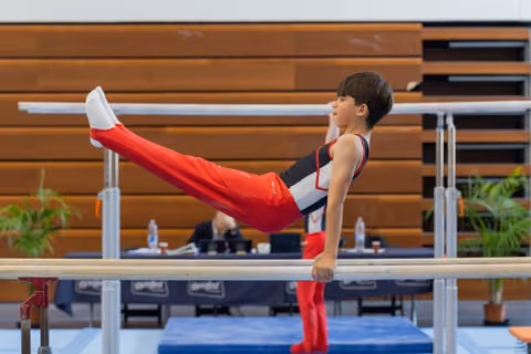 Focused young male gymnast holds a straddle L-sit on parallel bars, legs extended, expression intent during a meet.