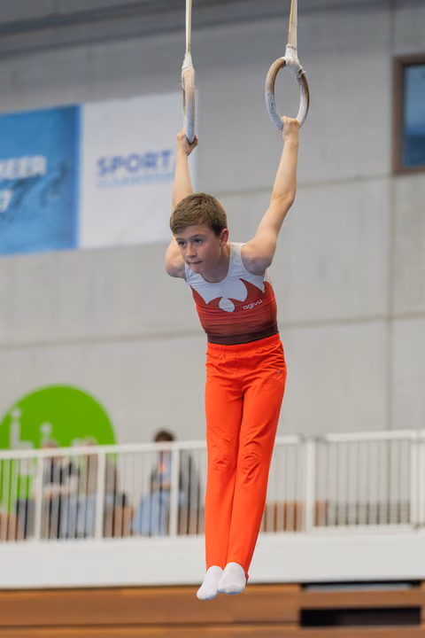 Young male gymnast hangs from still rings with focused expression, wearing red and white leotard in indoor gymnasium.