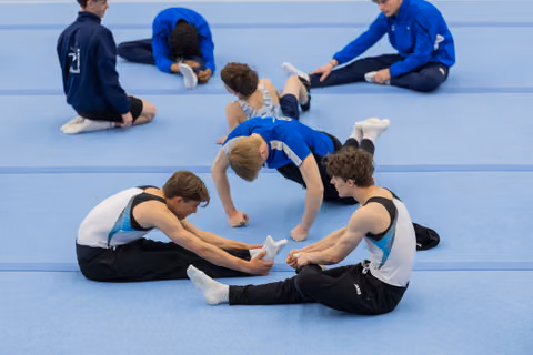 Young male gymnasts stretch and assist each other on a blue gymnasium floor, relaxed and focused during warm-up.