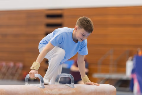 Young gymnast grips pommel horse handles with wrist guards, leaning forward with concentration during a gymnastics event.