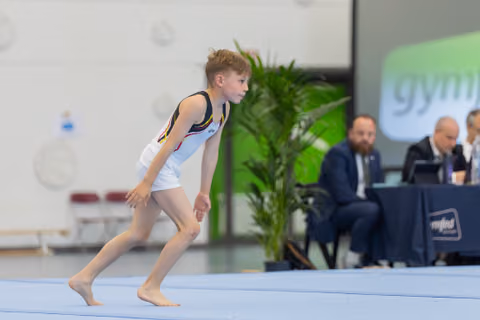 Young male gymnast crouches into a focused run-up stance on the floor exercise mat, judges seated behind.
