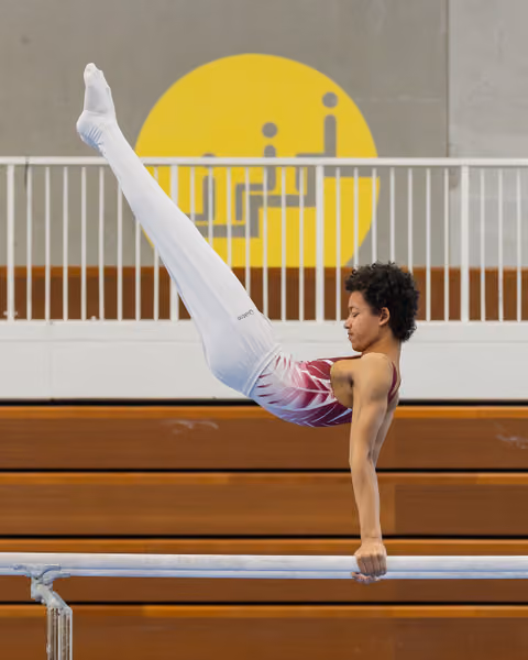 Young male gymnast holds a powerful pike position on parallel bars, legs raised high, expression intense and focused.