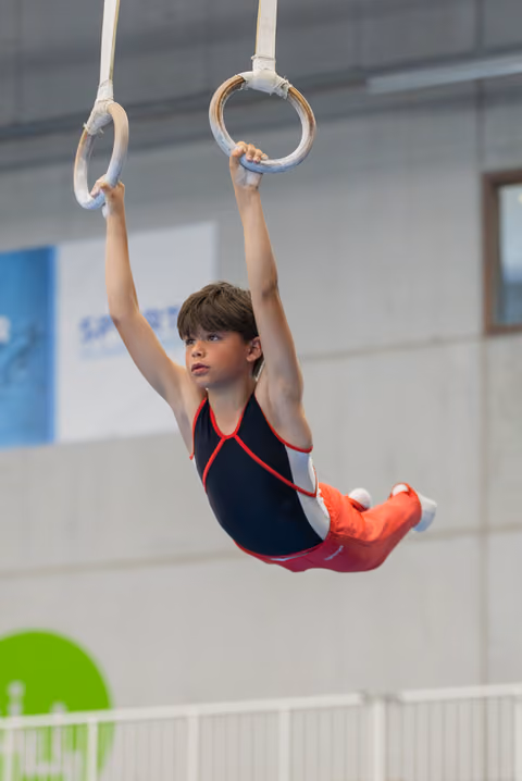 Focused young male gymnast hangs from still rings in a navy and red leotard during an indoor gymnastics competition.