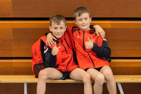 Two young boys in red sports jackets sit on a bench, arms around each other, both giving enthusiastic thumbs up and smiling.