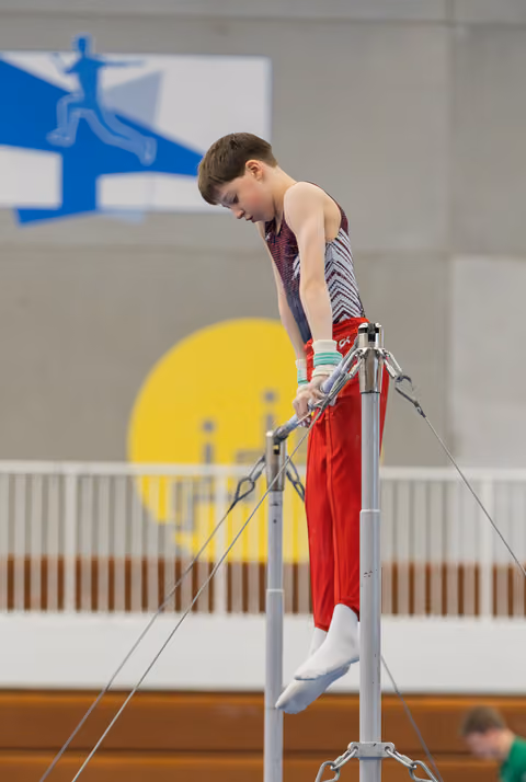 Young male gymnast grips the high bar with intense focus, body angled downward in a gymnastics hall.