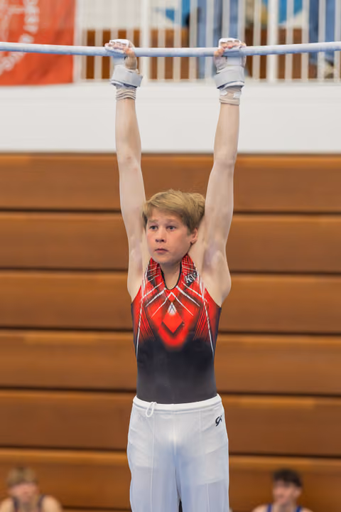 Young male gymnast hangs from the high bar with focused concentration, wearing a red and black leotard in a gymnasium.