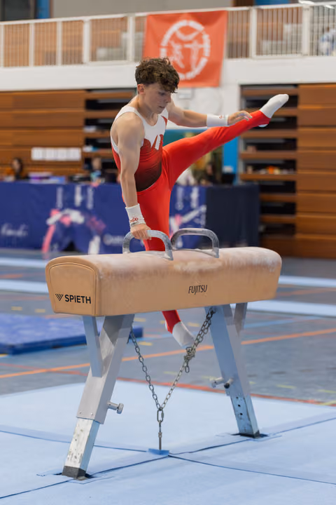 Young male gymnast performs on pommel horse, leg raised high, focused expression, indoor gymnastics hall.