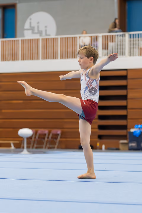 Focused young male gymnast performs a leg raise on the floor exercise, arms outstretched for balance in an indoor gym.