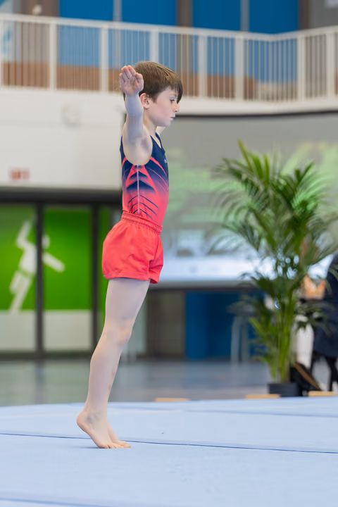 Young male gymnast stands on tiptoes on the floor mat, arm raised, focused expression during a gymnastics event.