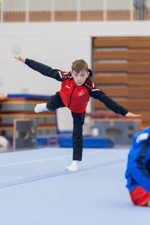 Young boy in red vest balances on one leg, arms outstretched, focused expression during floor gymnastics routine.